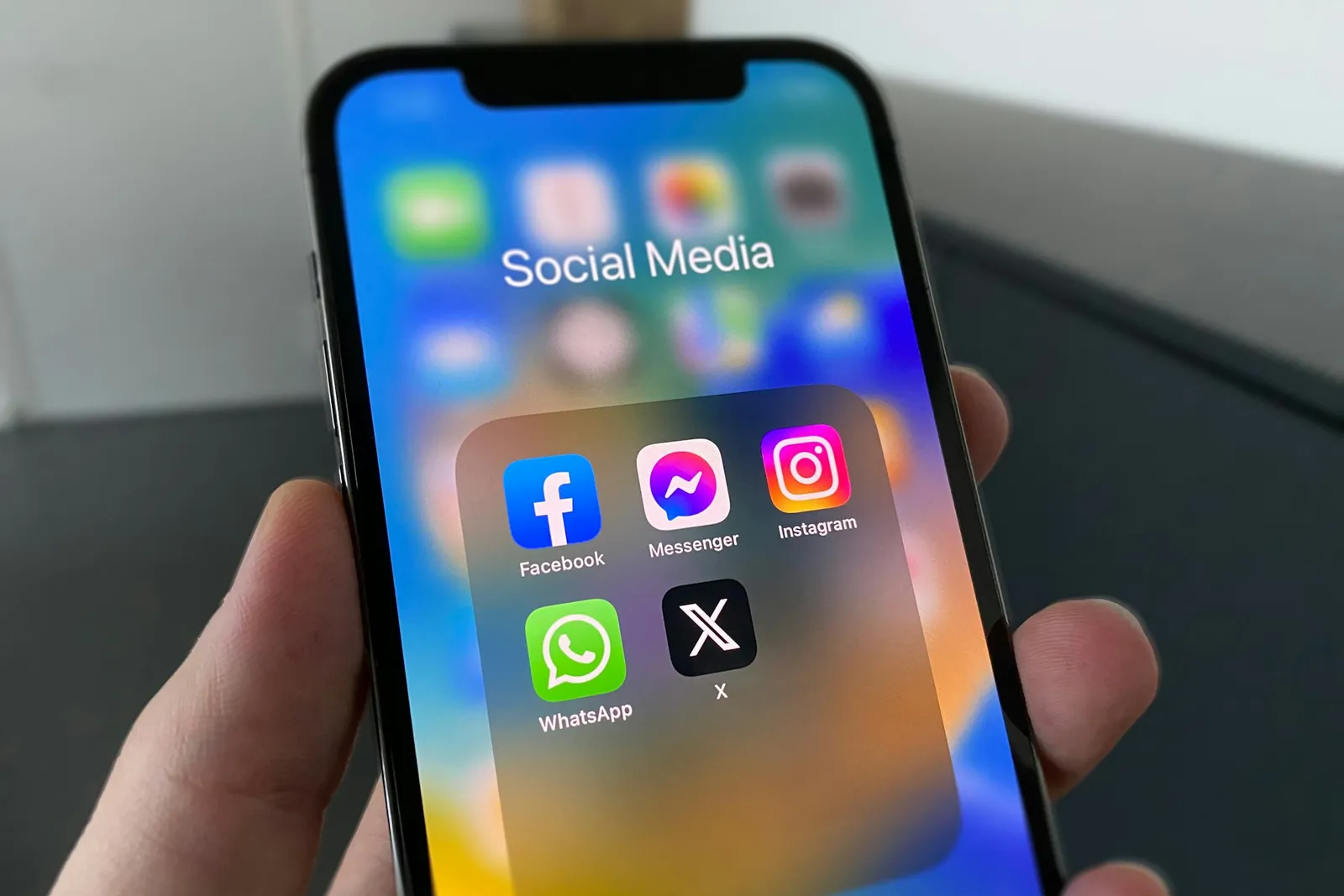 A hand holding a phone with a Social Media folder open: Facebook, Messenger, Instagram, WhatsApp, X.